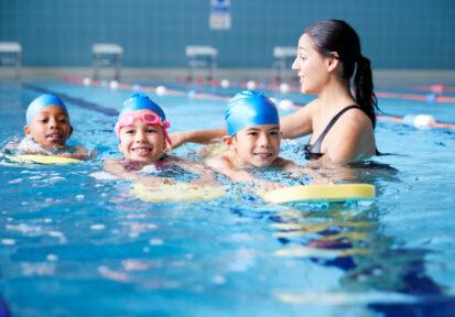 Female Coach In Water Giving Group Of Children Swimming Lesson In Indoor Pool